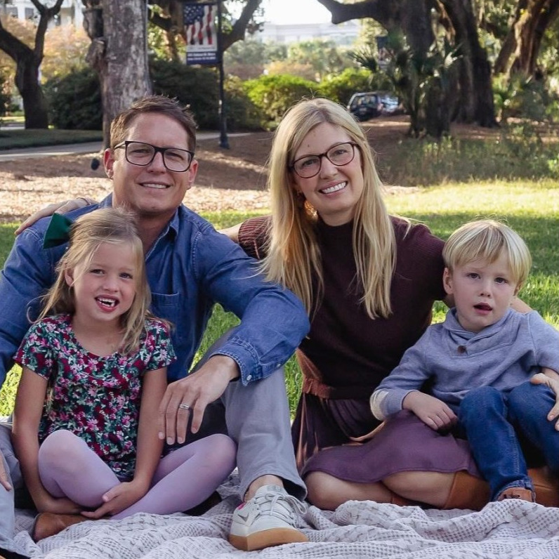 Clay Estabrooke with his family, sitting on a blanket on a park lawn under live oaks