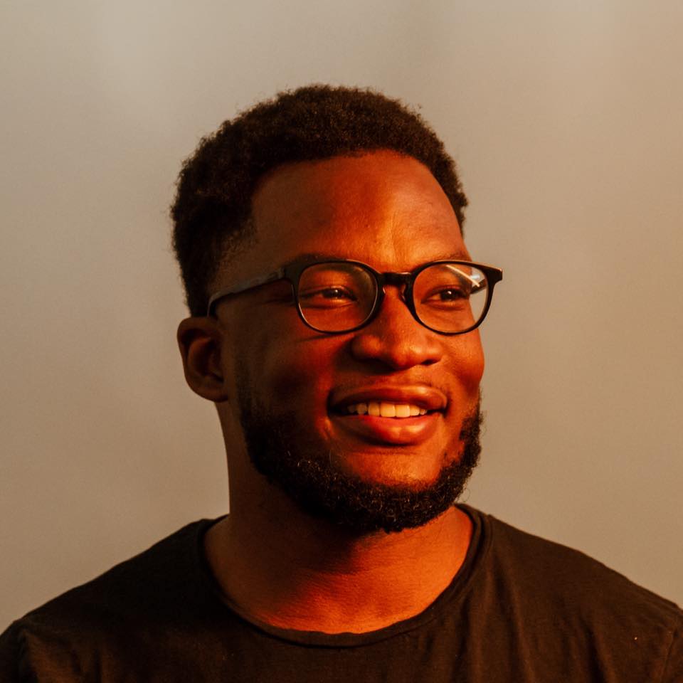 Harrison Wheeler in a black t-shirt and dark-rimmed glasses, smiling, lit warm against a pale wall