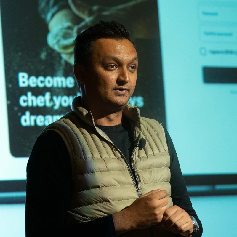 Ishan Singh speaking on stage in a tan vest, an SRVE slide projected behind him