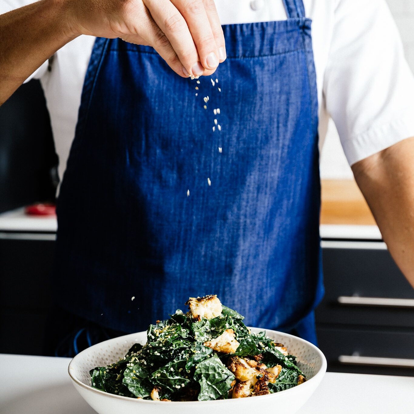 A chef in a blue denim apron scattering seeds over a bowl of kale and roasted cauliflower