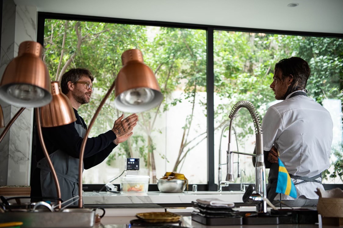 Two chefs talking across a kitchen island set up for service, garden beyond the window