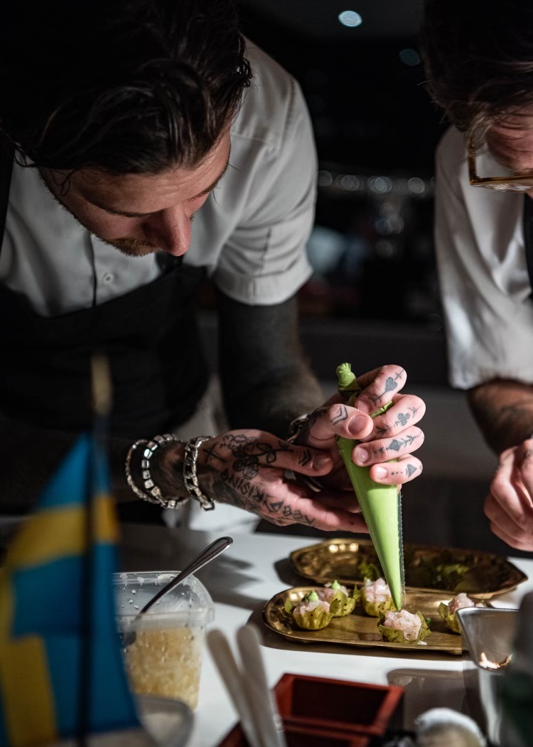 A chef piping garnish onto canapés on brass trays at a pop-up service