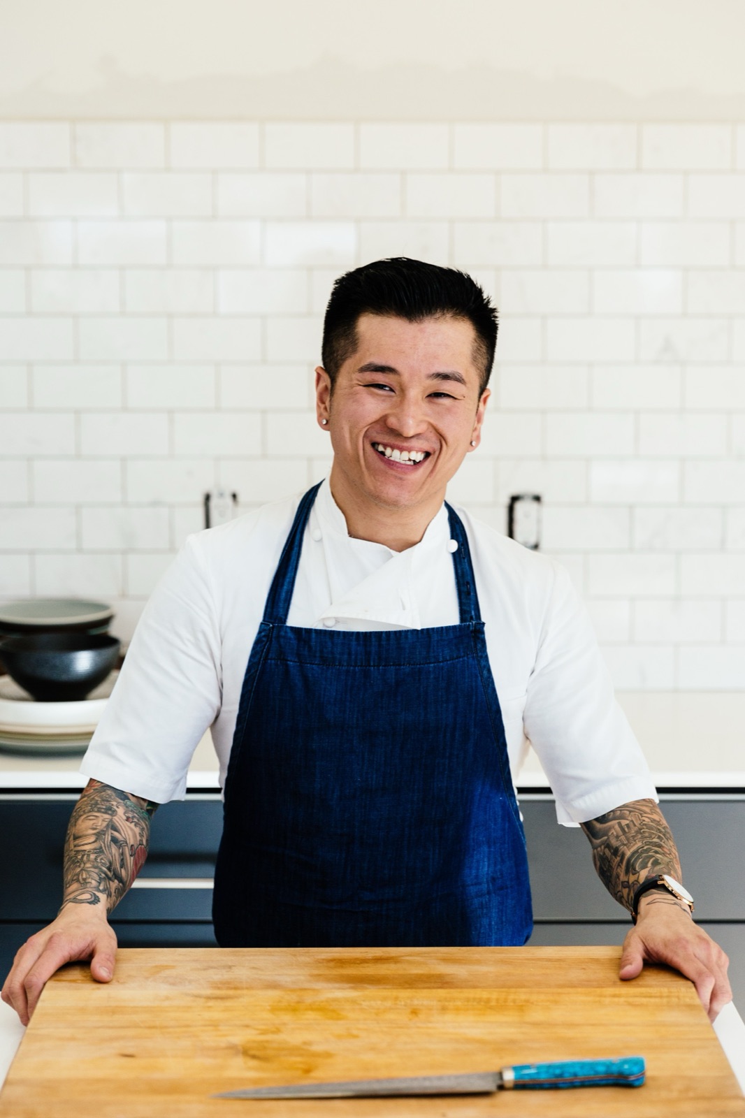 A smiling chef in a blue denim apron standing behind a wooden cutting board