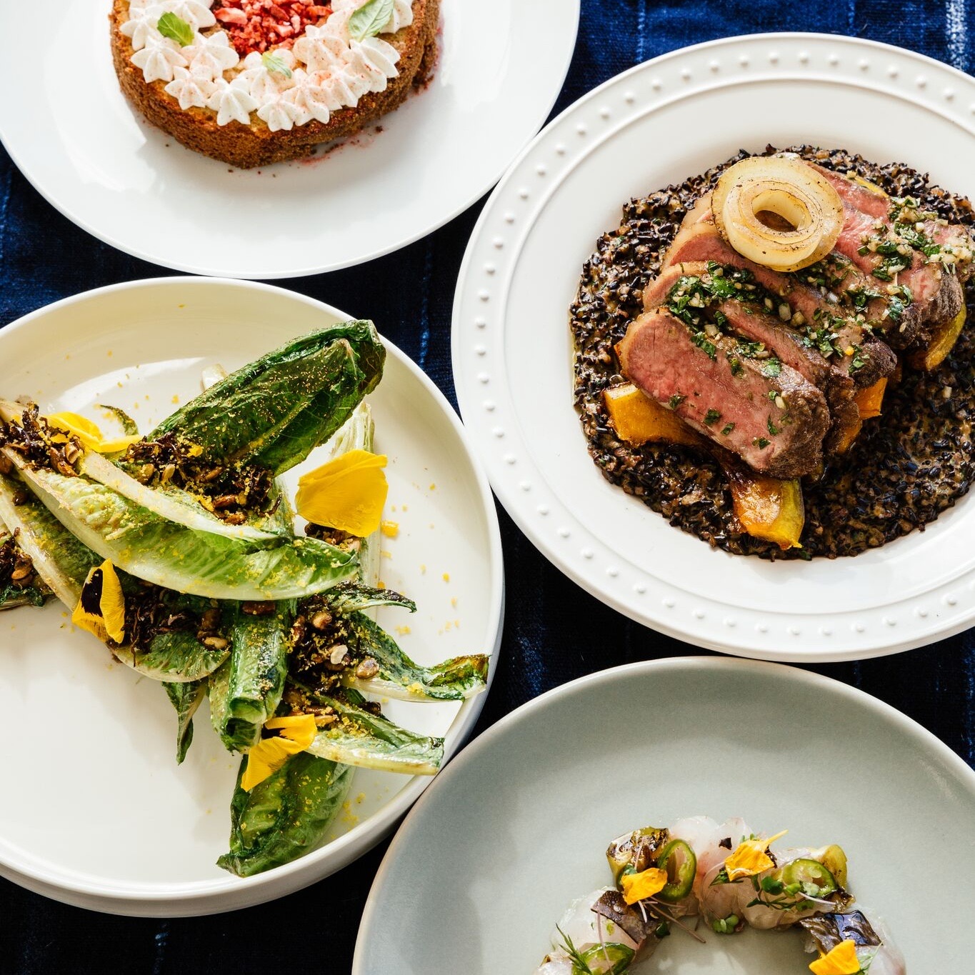 Four plated courses on a dark blue cloth: a cream-piped cake, sliced steak with chimichurri over wild rice, charred romaine with seeds and petals, and a crudo with jalapeño