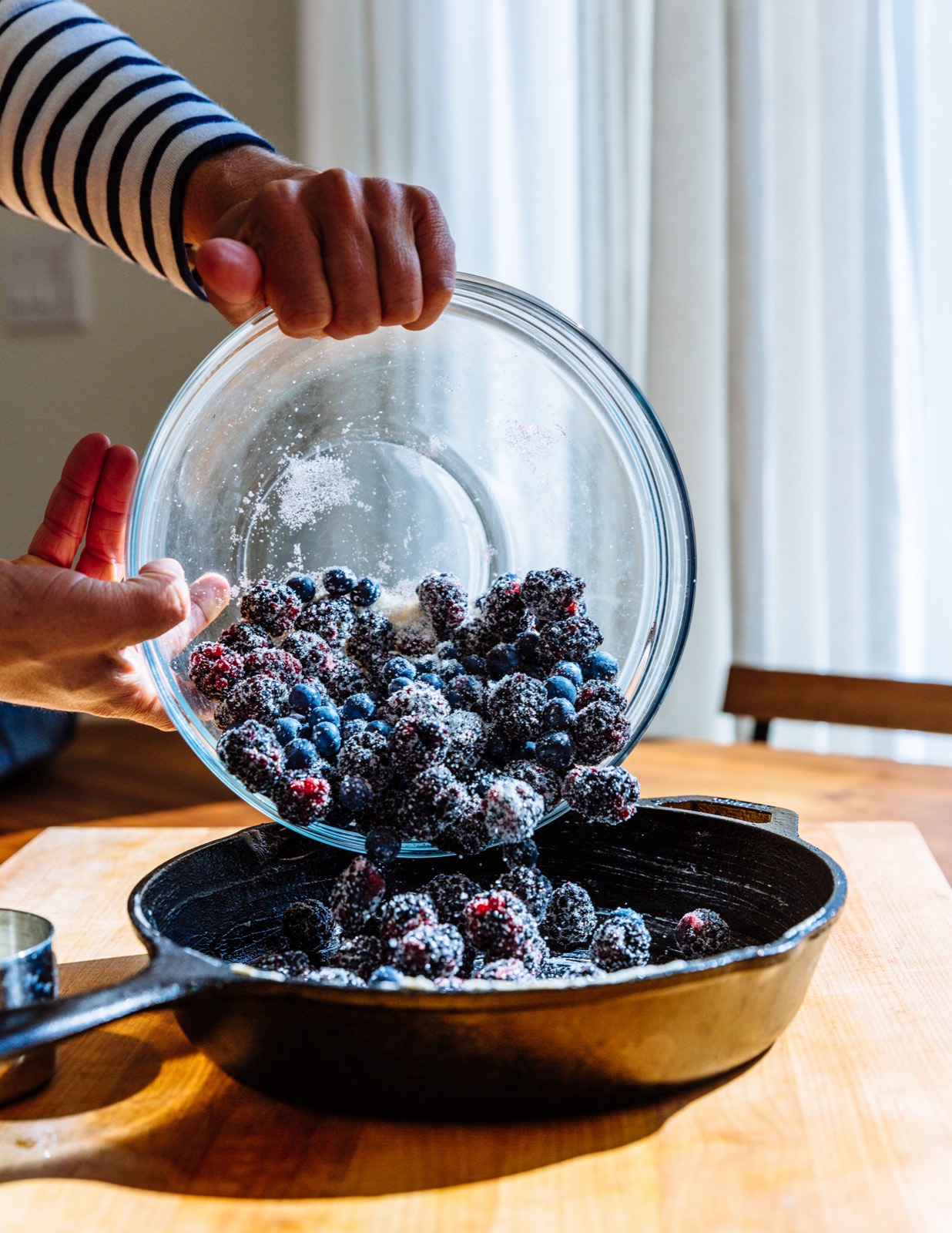 Sugared blackberries and blueberries being tipped from a glass bowl into a cast-iron skillet on a kitchen table