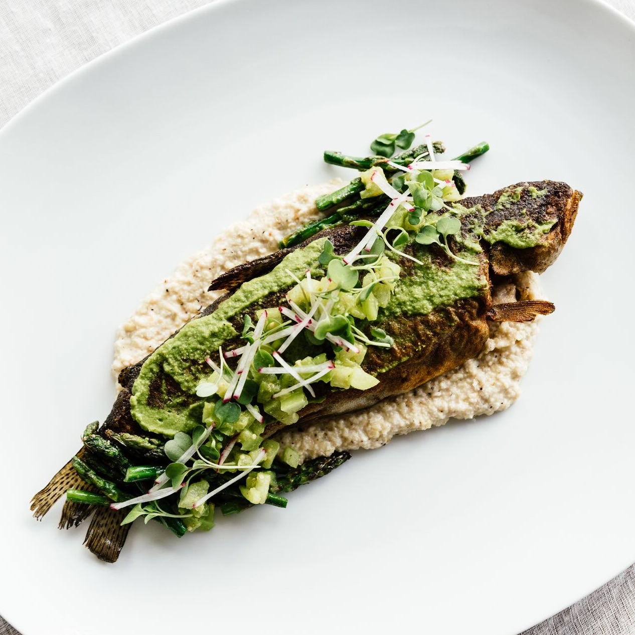 A whole roasted fish on grits with salsa verde, charred asparagus, and radish sprouts, plated on a wide white dish