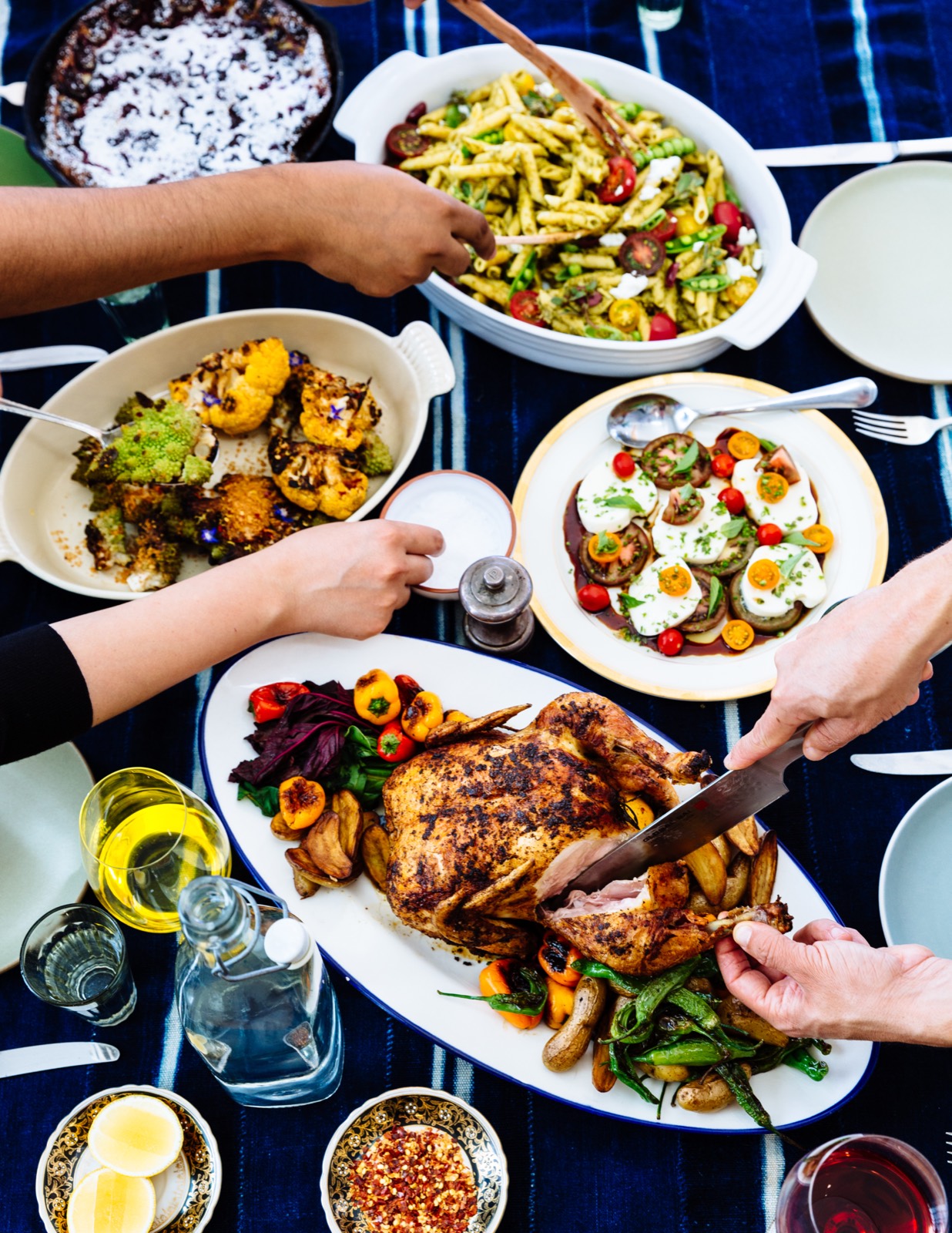 An overhead table of shared dishes — roast chicken being carved, pesto pasta salad, roasted cauliflower, and a tomato and mozzarella plate — with several hands reaching in
