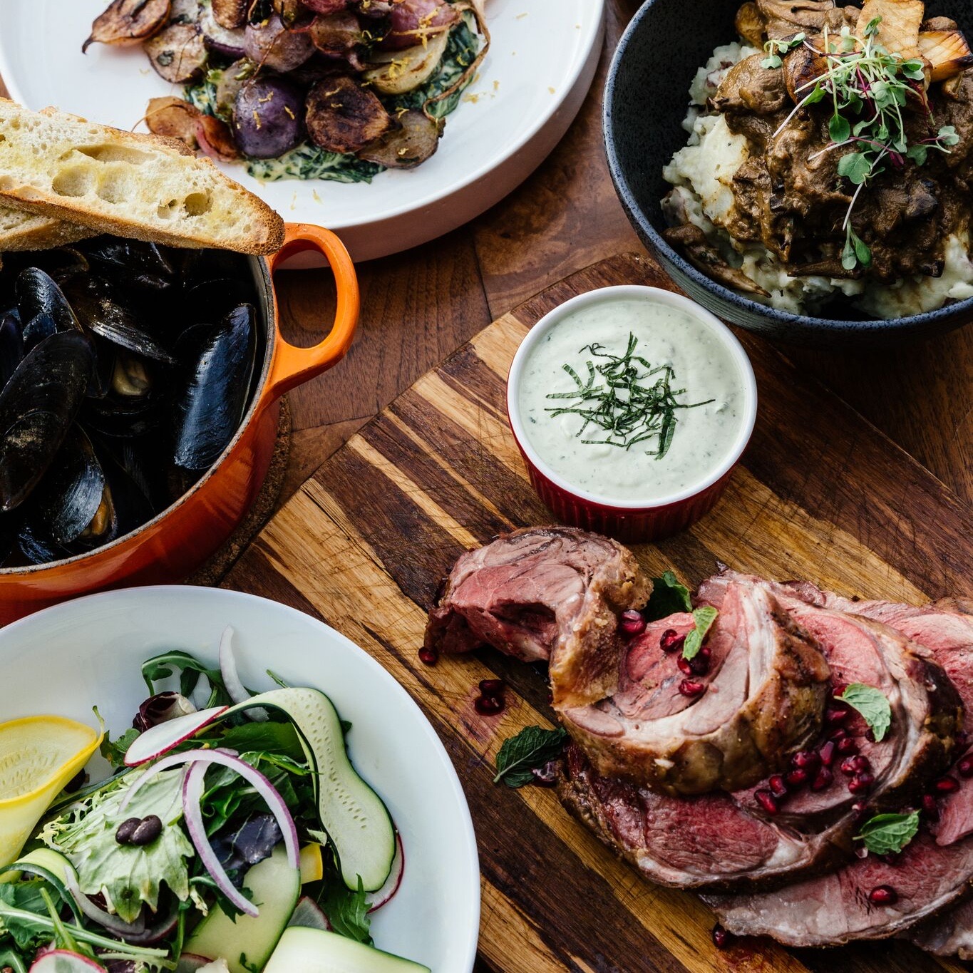 An overhead spread on a wooden table: a pot of mussels with toasted bread, roast potatoes over greens, mushroom stroganoff on mash, a green salad, and sliced rack of lamb with pomegranate