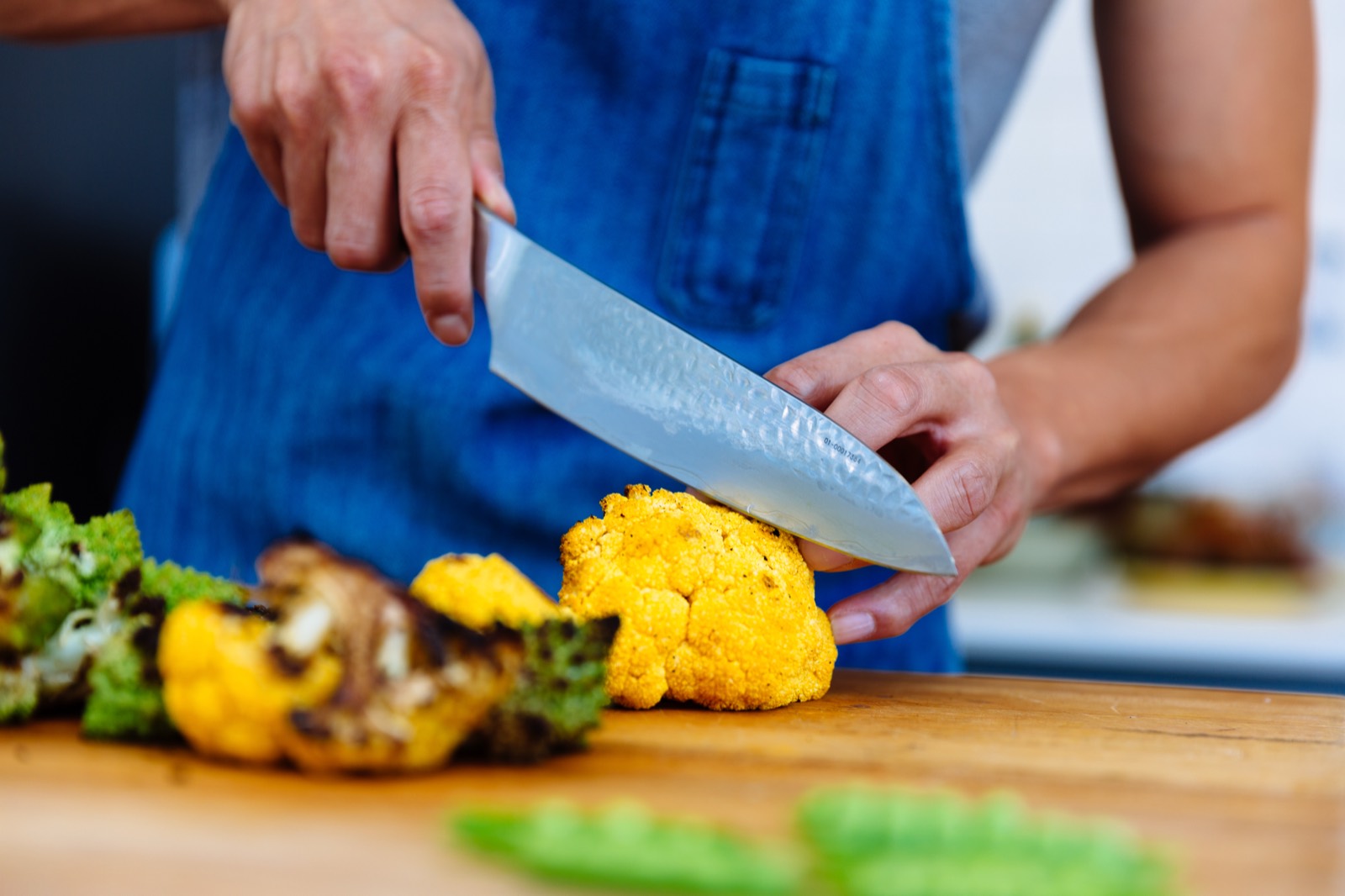 A cook in a denim apron slicing a head of roasted yellow cauliflower on a wooden board