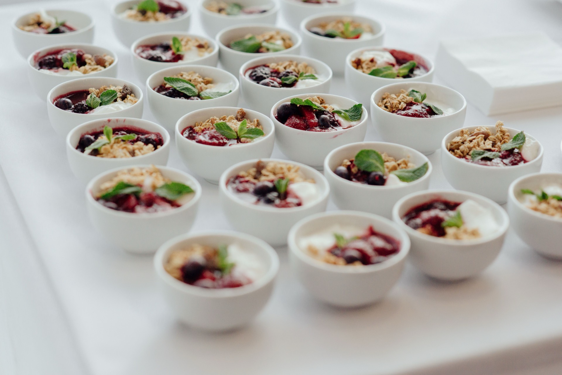 Rows of small white bowls plated identically with yoghurt, berry compote, granola, and a mint leaf, laid out on a linen table