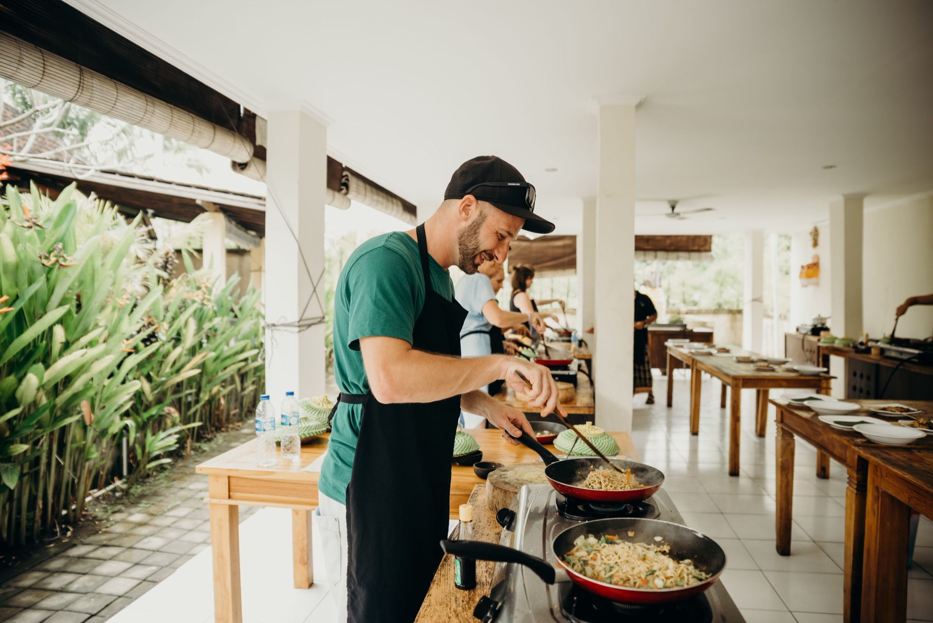 A student in an apron tossing noodles in a pan at an open-air cooking class, with more stations working down the counter behind them