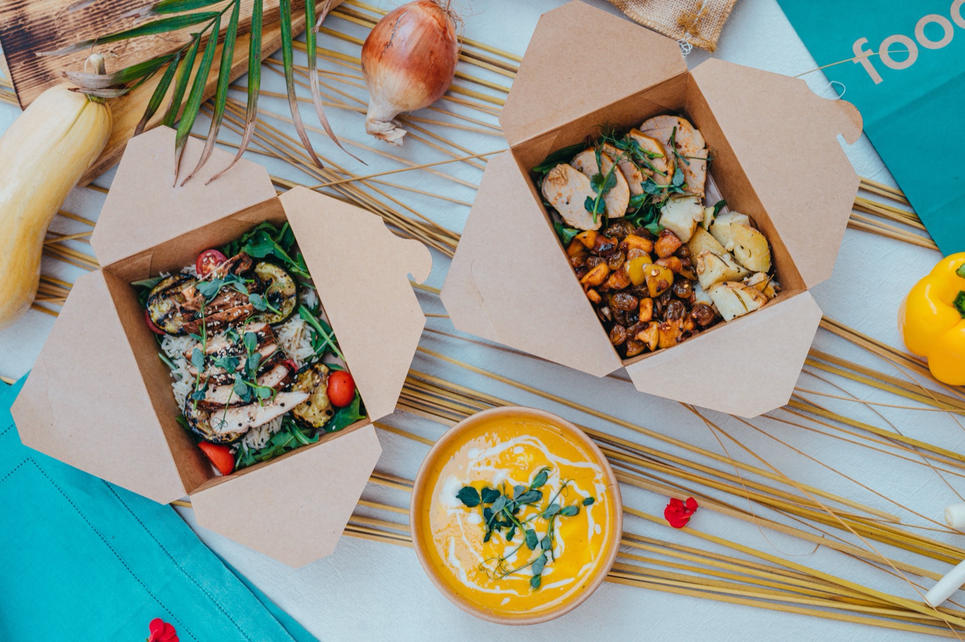 Two open kraft takeaway boxes — grilled chicken over rice with courgette, and roast squash with halloumi — beside a bowl of squash soup