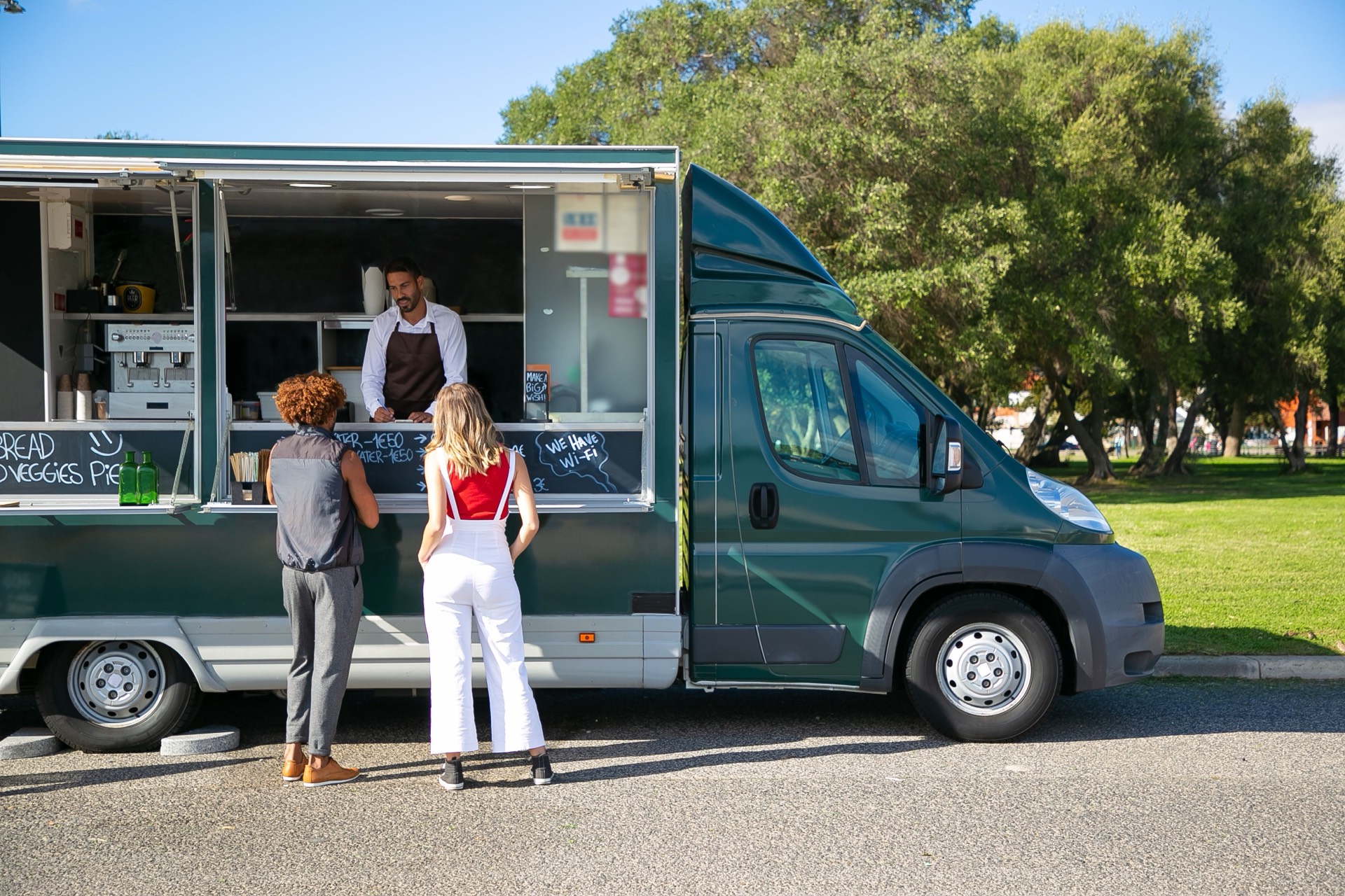 Two customers ordering at the window of a green food truck parked by a park, the cook taking their order behind a chalkboard menu