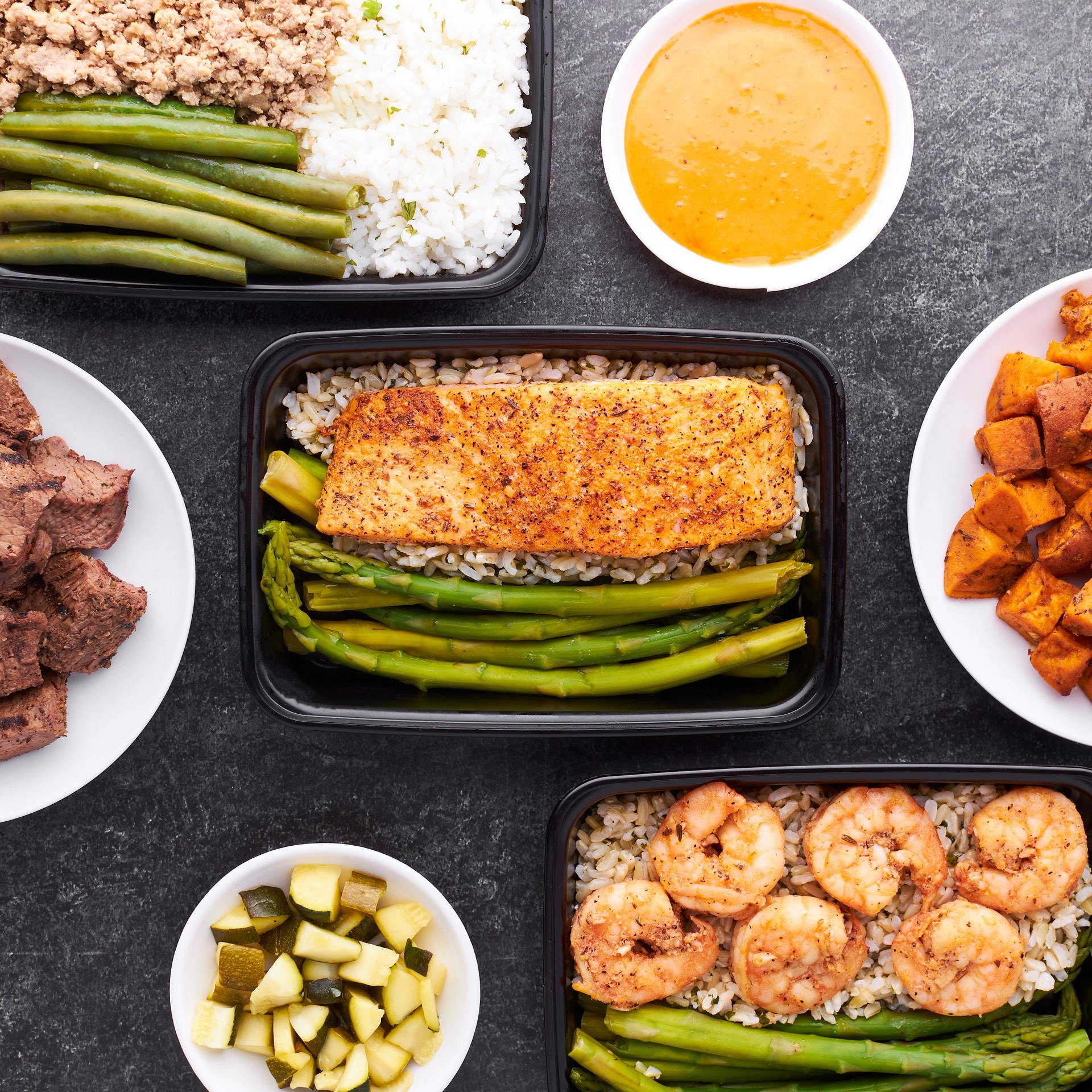 Portioned meal containers on a slate counter: salmon and asparagus over rice, shrimp and brown rice, turkey with green beans, and side bowls of sweet potato, steak, and squash