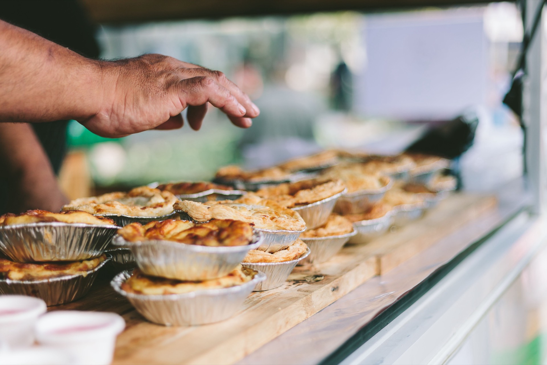 A cook reaching across a market counter lined with freshly baked pies in foil tins