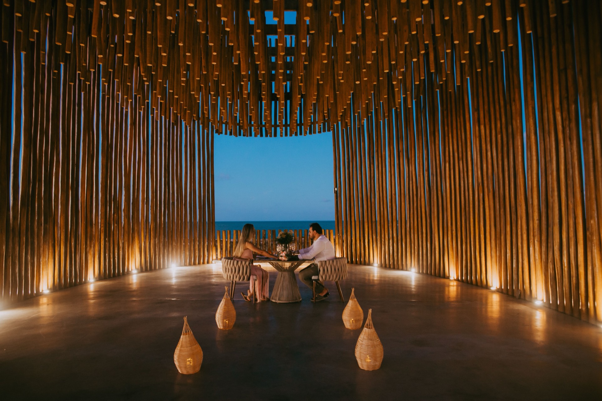 Two guests at a candlelit table for two inside a timber pavilion, lanterns on the floor and the ocean framed beyond
