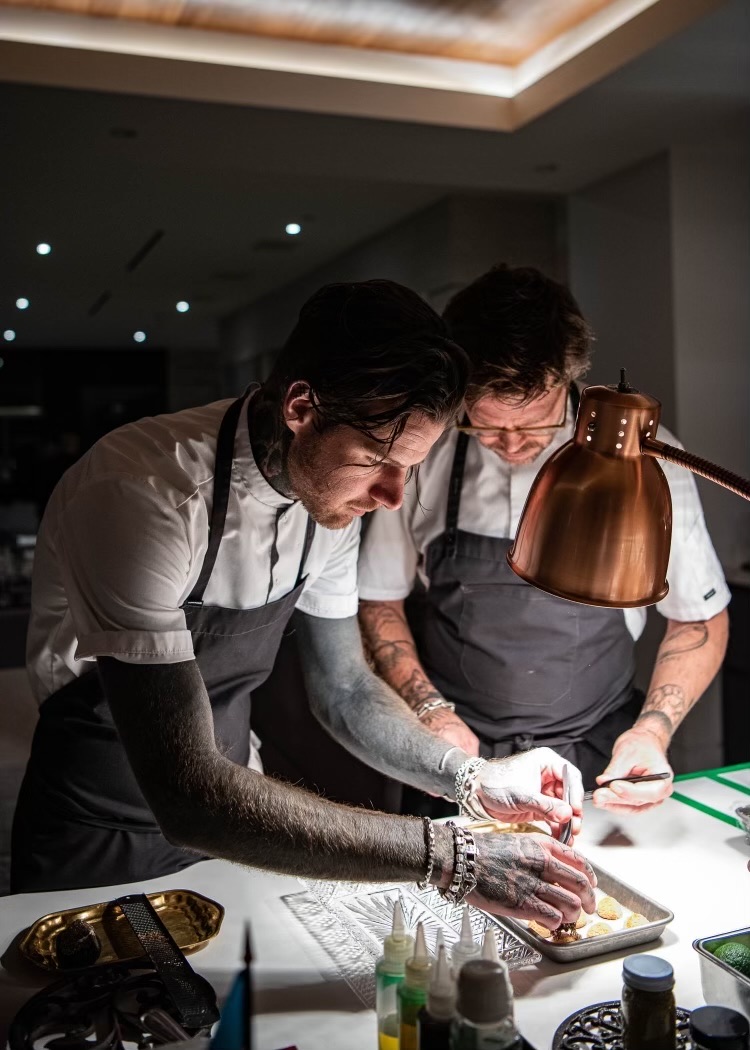 Two chefs plating canapés under a copper heat lamp